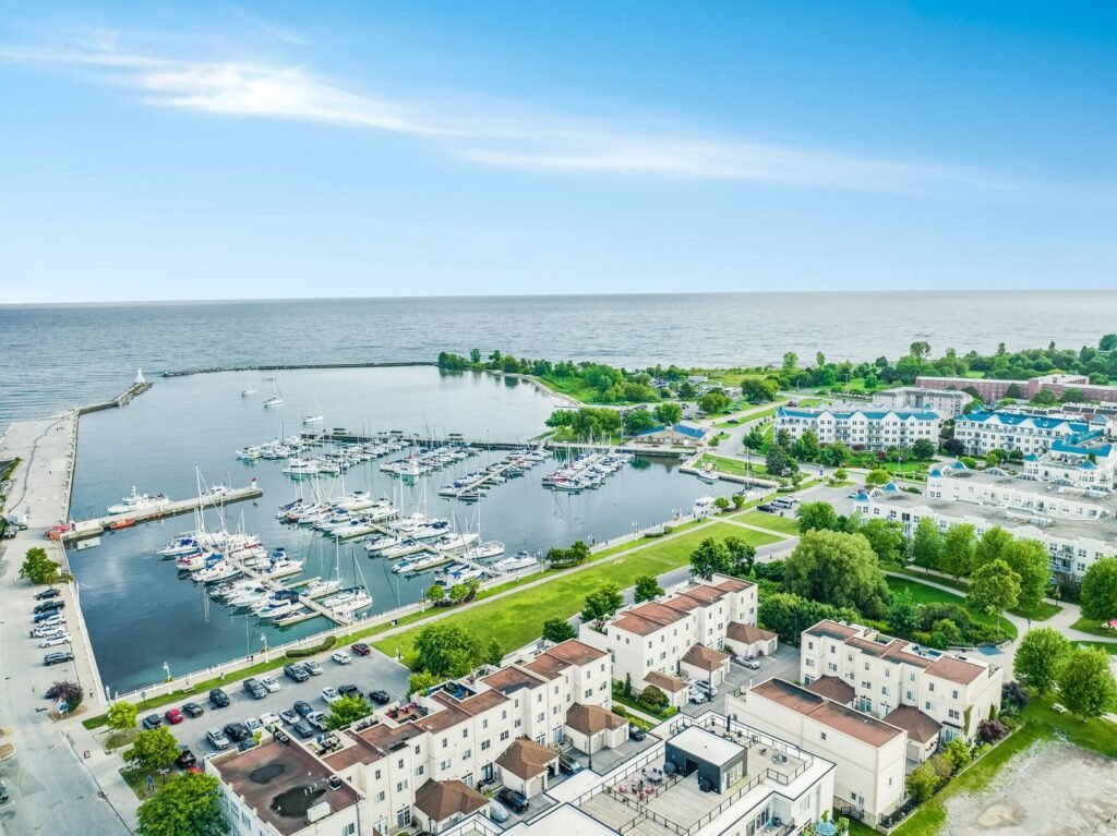 Aerial view of a marina with docked boats, nearby buildings, green spaces, and a large body of water under a clear blue sky.