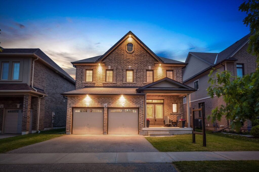 A two-story brick house with two garage doors, illuminated exterior lights, a front porch, and a small lawn at dusk.
