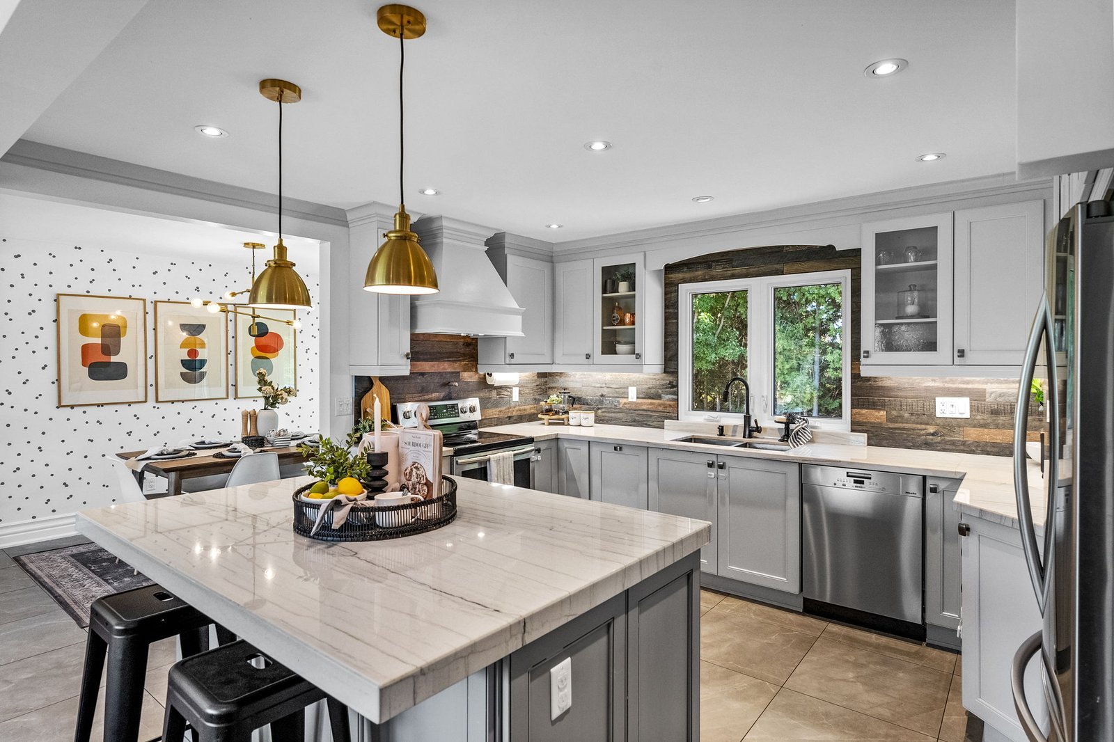 Modern kitchen with white cabinets, stainless steel appliances, marble countertop island, gold pendant lights, and a dining area with three framed prints on a polka-dot accent wall.