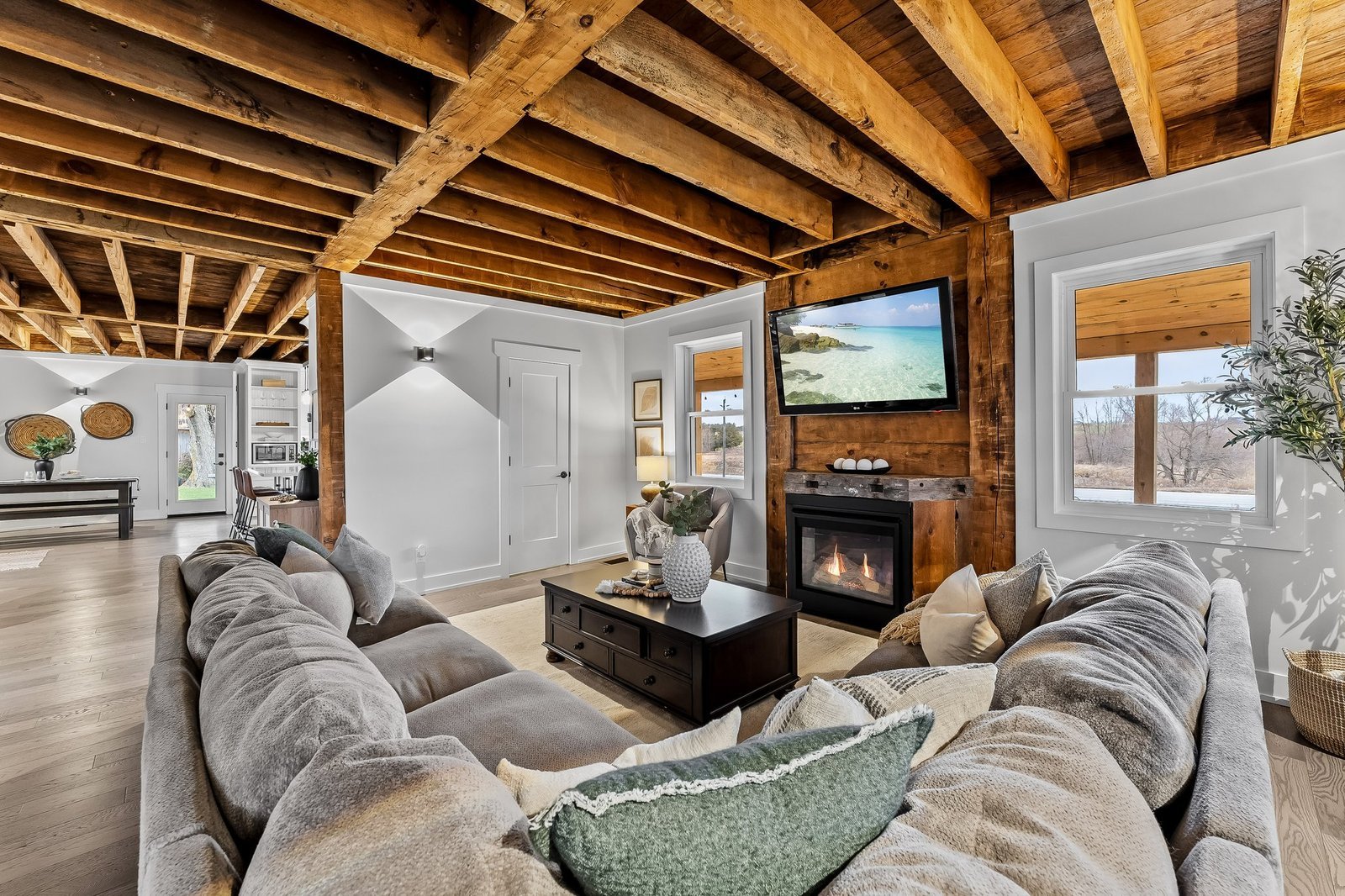 A cozy living room with a gray sectional sofa, wooden ceiling beams, a fireplace, wall-mounted TV, and large windows letting in natural light.