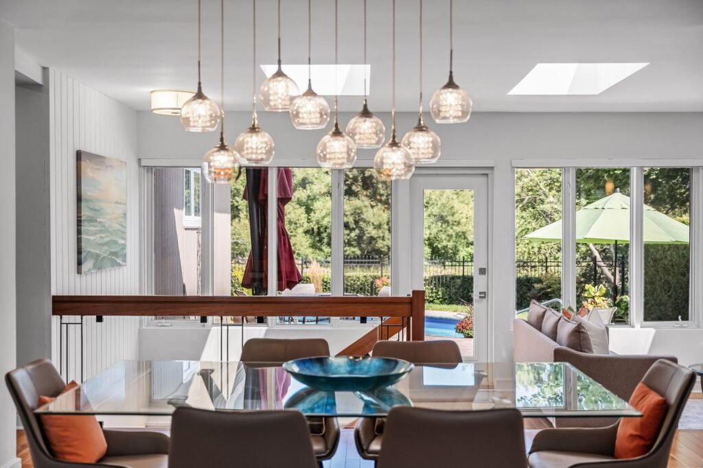 Modern dining area with glass table, contemporary pendant lights, and a view of a backyard patio through large windows and glass doors.
