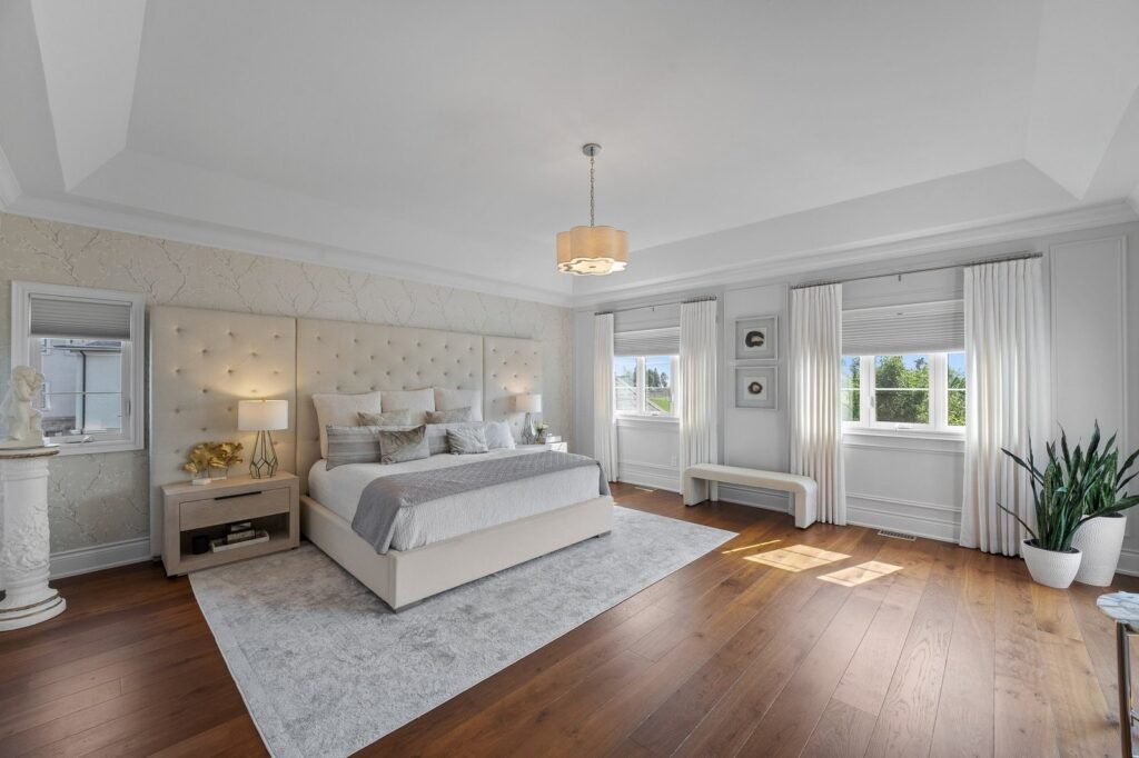 Spacious bedroom with a large bed, two nightstands, a white bench, hardwood floors, large windows with white curtains, and potted plants.