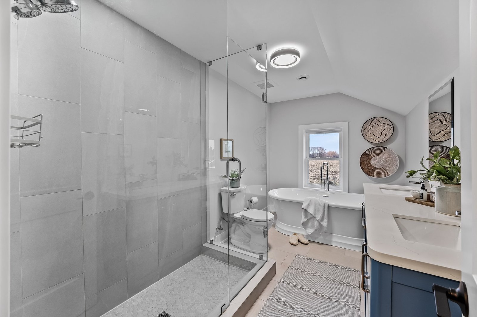 Modern bathroom with large glass shower, freestanding bathtub by a window, toilet, double sink vanity, and neutral decor accents.