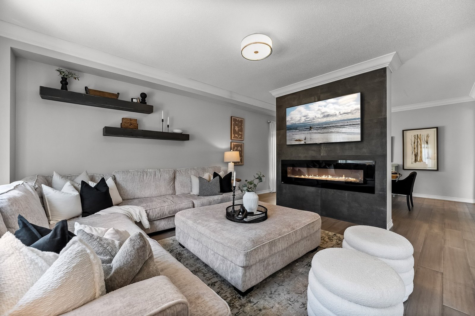 Modern living room with a large sectional sofa, ottoman, wall-mounted fireplace, and TV. Neutral tones and minimalist decor create a cozy and contemporary atmosphere.