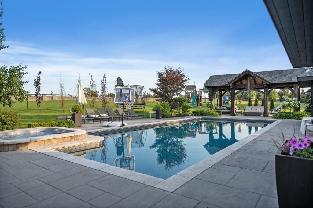 Outdoor swimming pool with adjacent hot tub, lounge chairs, a basketball hoop in the pool, and a wooden pavilion set in a landscaped backyard under a clear blue sky.