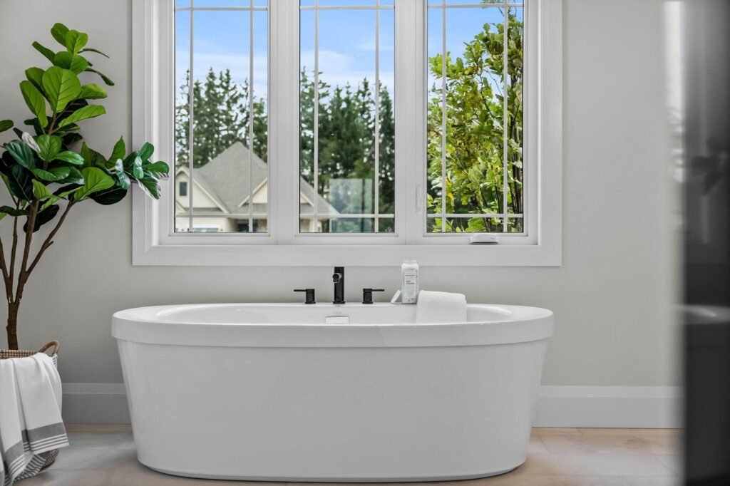 A white freestanding bathtub sits below large windows with a view of trees and a house. A towel and plant are nearby, giving the bathroom a bright and clean look.