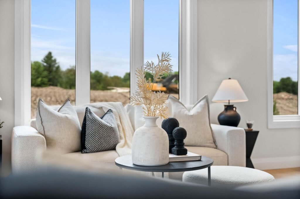A modern living room with a white sofa, neutral pillows, a small round table with decorative objects, and large windows overlooking greenery.