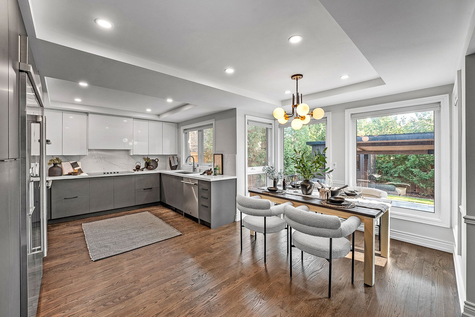Modern kitchen and dining area with gray cabinets, wooden floors, a dining table set for four, large windows, and contemporary lighting.