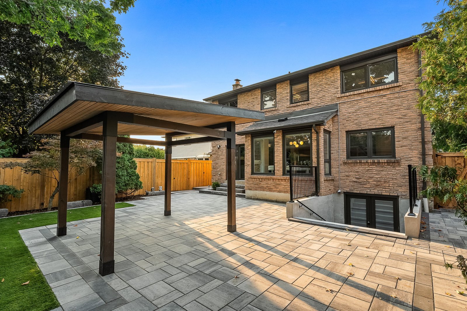 A two-story brick house with large windows, a covered patio structure, stone pavers, and a wooden fence surrounding a landscaped backyard.
