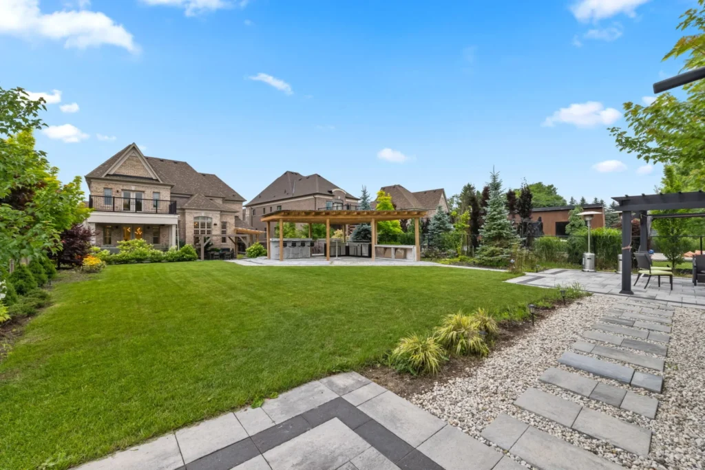 Spacious backyard with a large green lawn, modern gazebo, outdoor kitchen area, stone pathway, and upscale houses in the background under a blue sky.