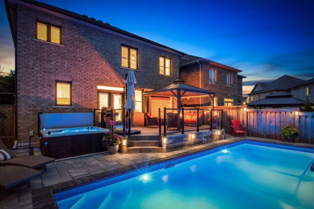 Backyard with illuminated swimming pool, hot tub, gazebo, seating area, and brick two-story house at dusk.