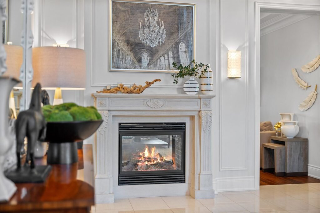 A lit fireplace with a stone mantel is centered below a framed artwork. Decorative items and a plant are on the mantel; a table with ornaments is in the foreground.