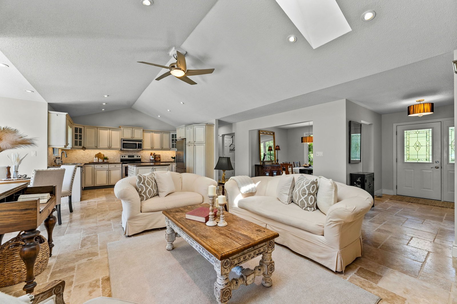 Open-concept living room and kitchen with neutral decor, beige tile flooring, white sofas, wooden coffee table, and modern ceiling fan under a vaulted ceiling with skylight.
