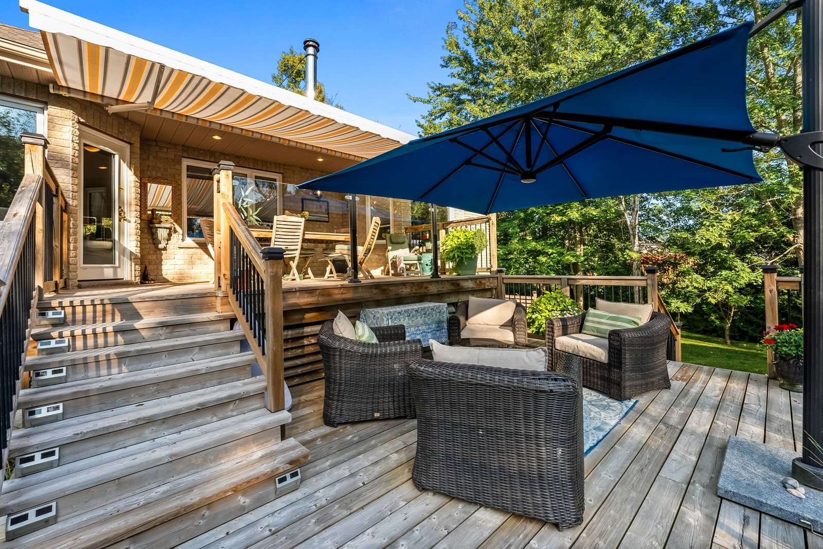 Wooden deck with wicker chairs, a blue patio umbrella, and stairs leading to a covered porch area; surrounded by trees and greenery.