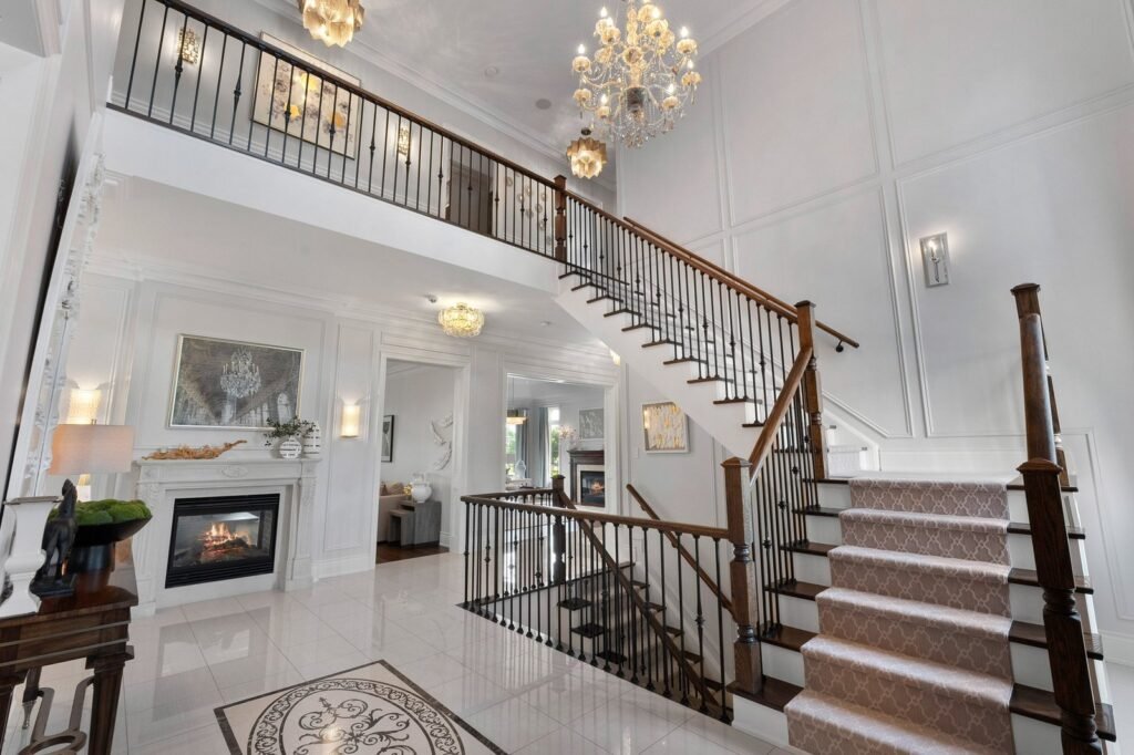 Modern, spacious foyer with a grand staircase, elegant chandeliers, a fireplace, tiled floors, and wall art. Upper and lower levels visible with natural light from windows.