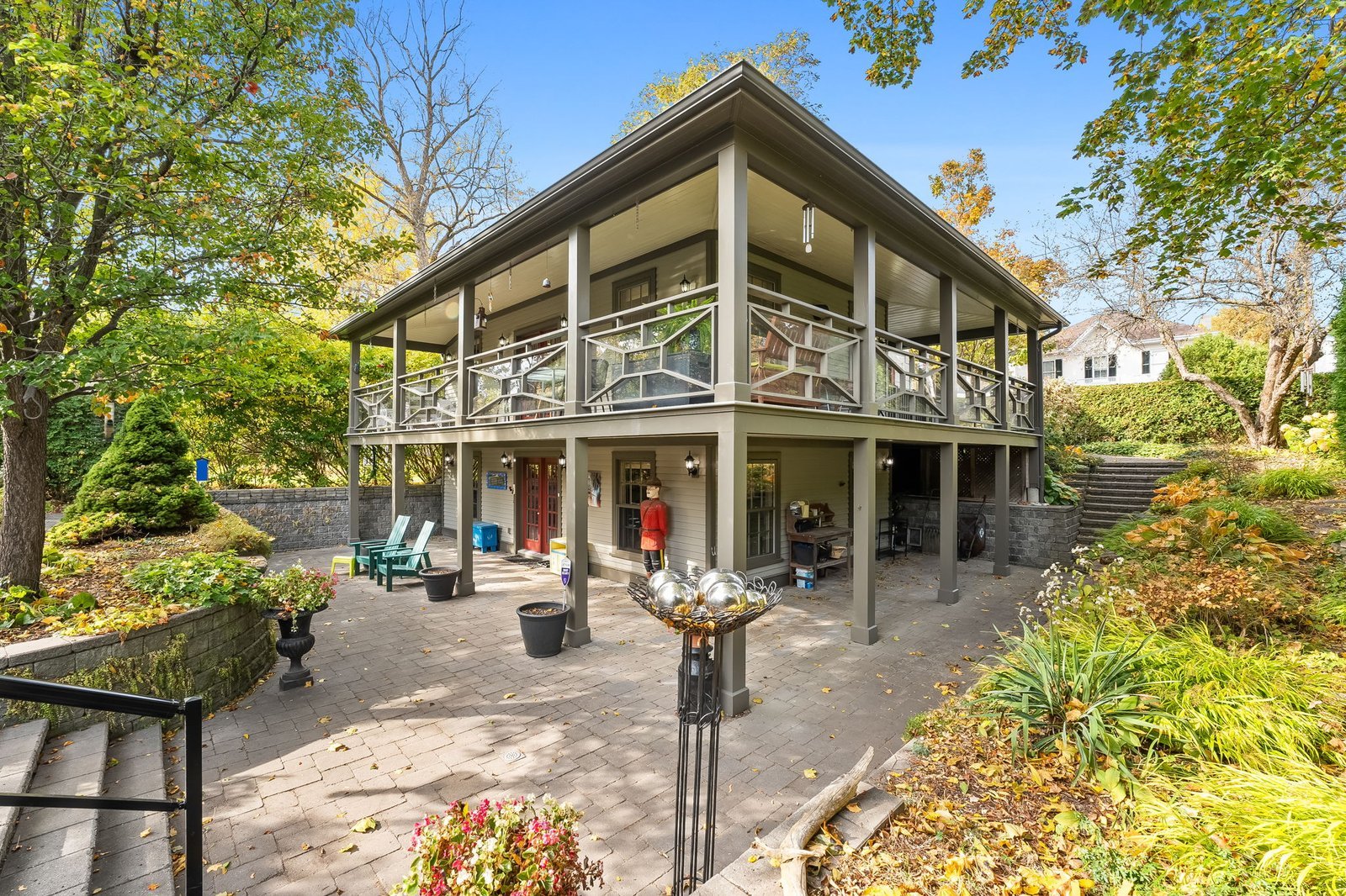 Two-story house with large covered balconies, glass railings, and an open ground floor patio, surrounded by trees and landscaped gardens on a sunny day.