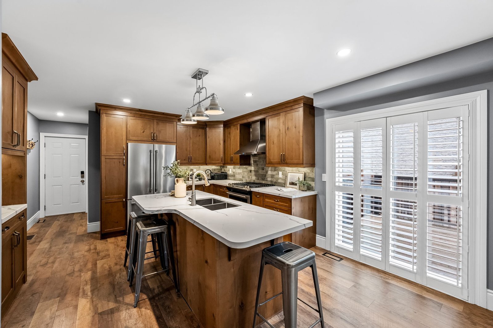 Modern kitchen with wood cabinets, stainless steel appliances, an island with bar stools, hardwood floors, and large windows with white plantation shutters.