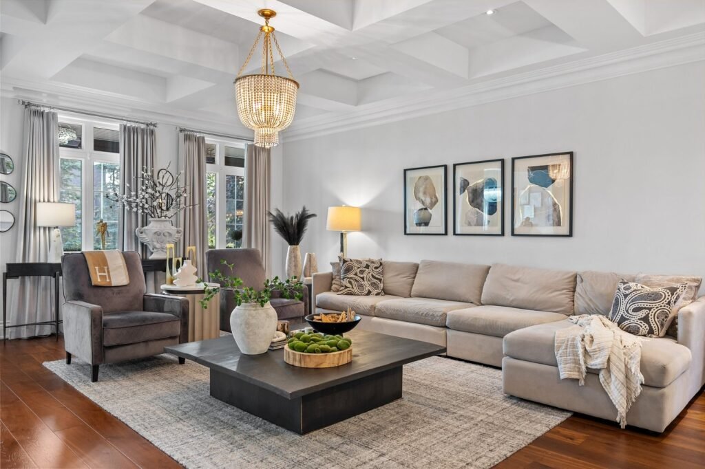A modern living room with a beige sectional sofa, two gray armchairs, a large black coffee table, wall art, and a chandelier on a coffered ceiling.