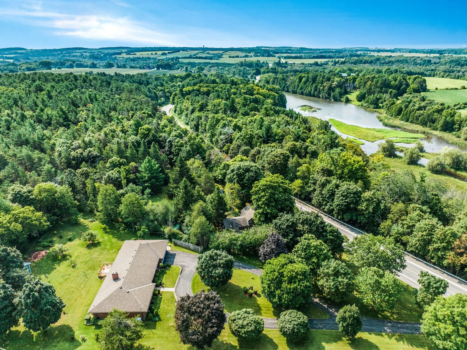 Aerial view of a large house surrounded by trees, open lawn, a winding road, and a river with green fields in the background under a clear blue sky.