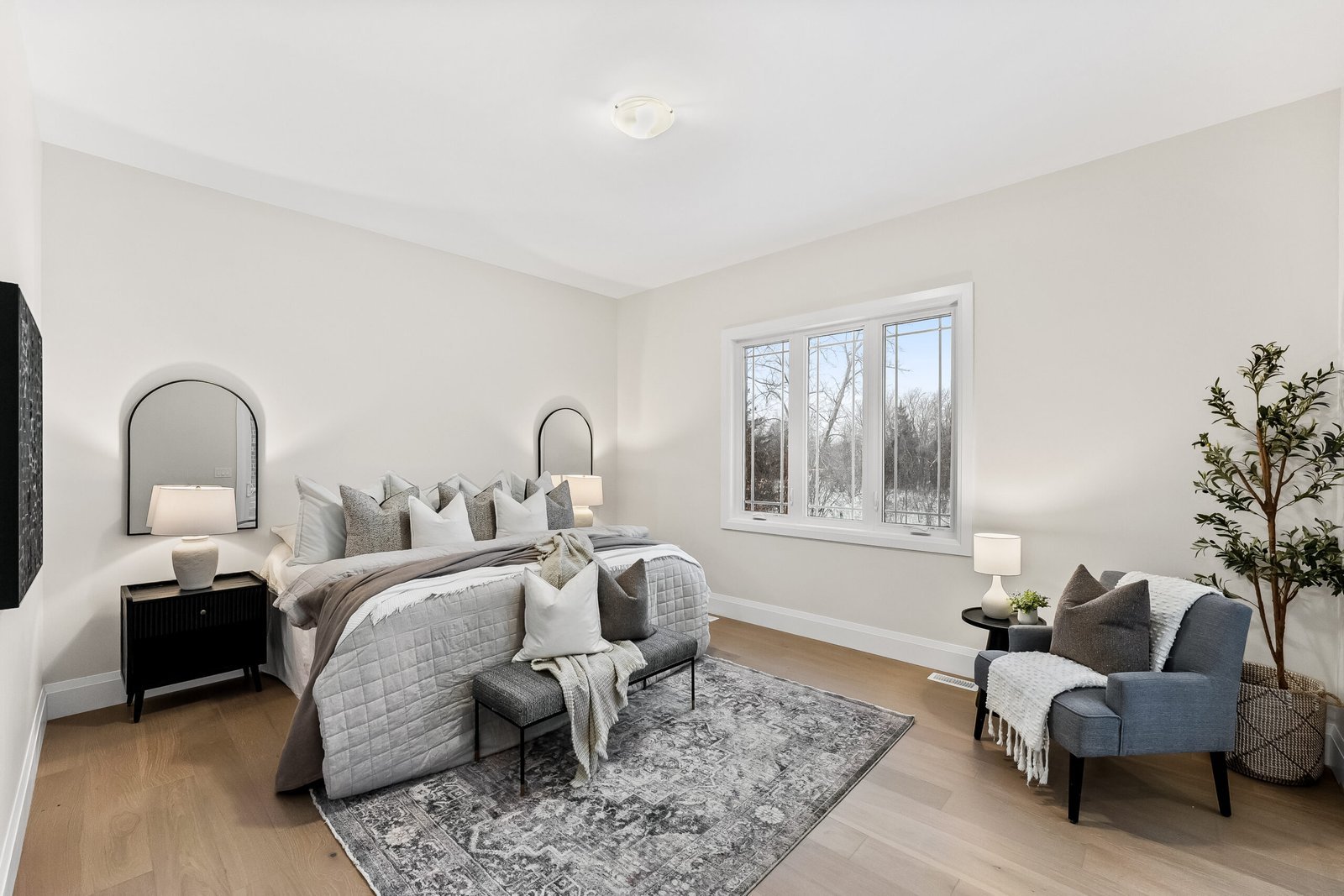 Modern bedroom with a large bed, decorative pillows, two nightstands with lamps, a patterned rug, an armchair with a blanket, potted plant, and a window with a view of trees.