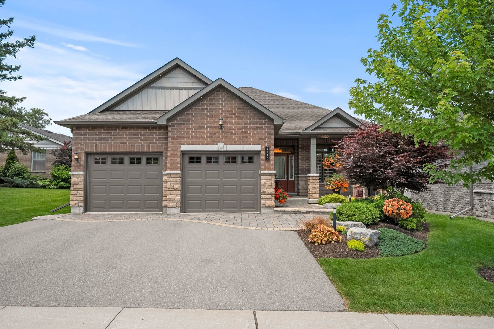 Single-story brick house with a double garage, landscaped front yard, green lawn, and various shrubs and flowers along the walkway.