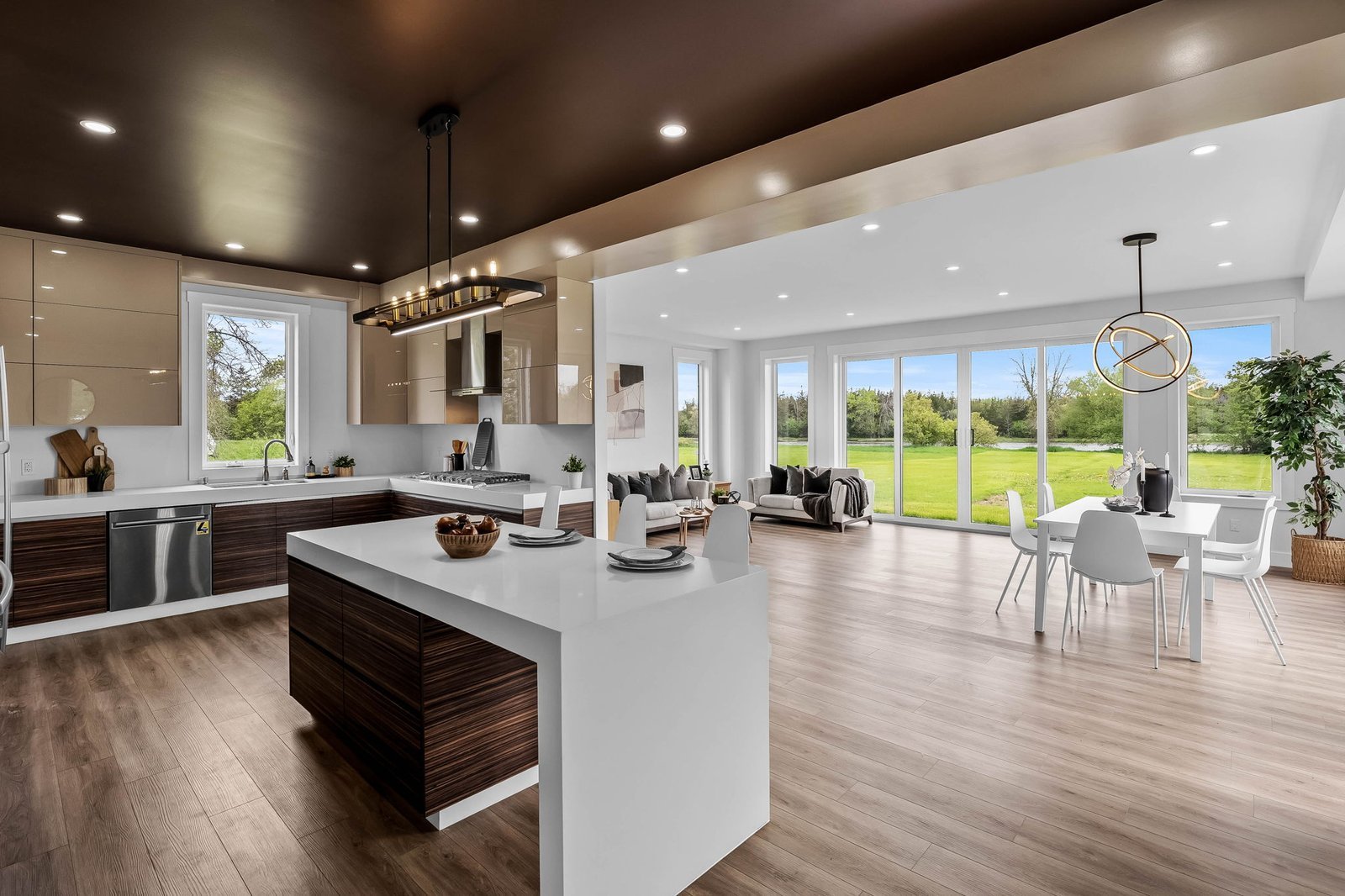 Modern open-concept kitchen and living area with wood floors, large island, contemporary lighting, and floor-to-ceiling windows overlooking a green lawn.
