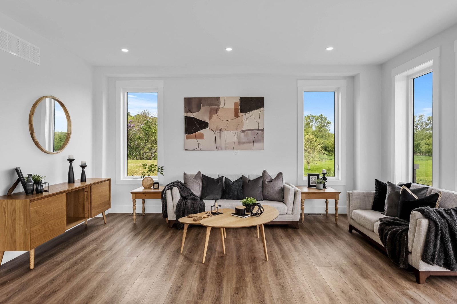 Modern living room with wood floors, large windows, a neutral sofa, armchair, abstract wall art, and a wooden coffee table. Greenery is visible outside the windows.