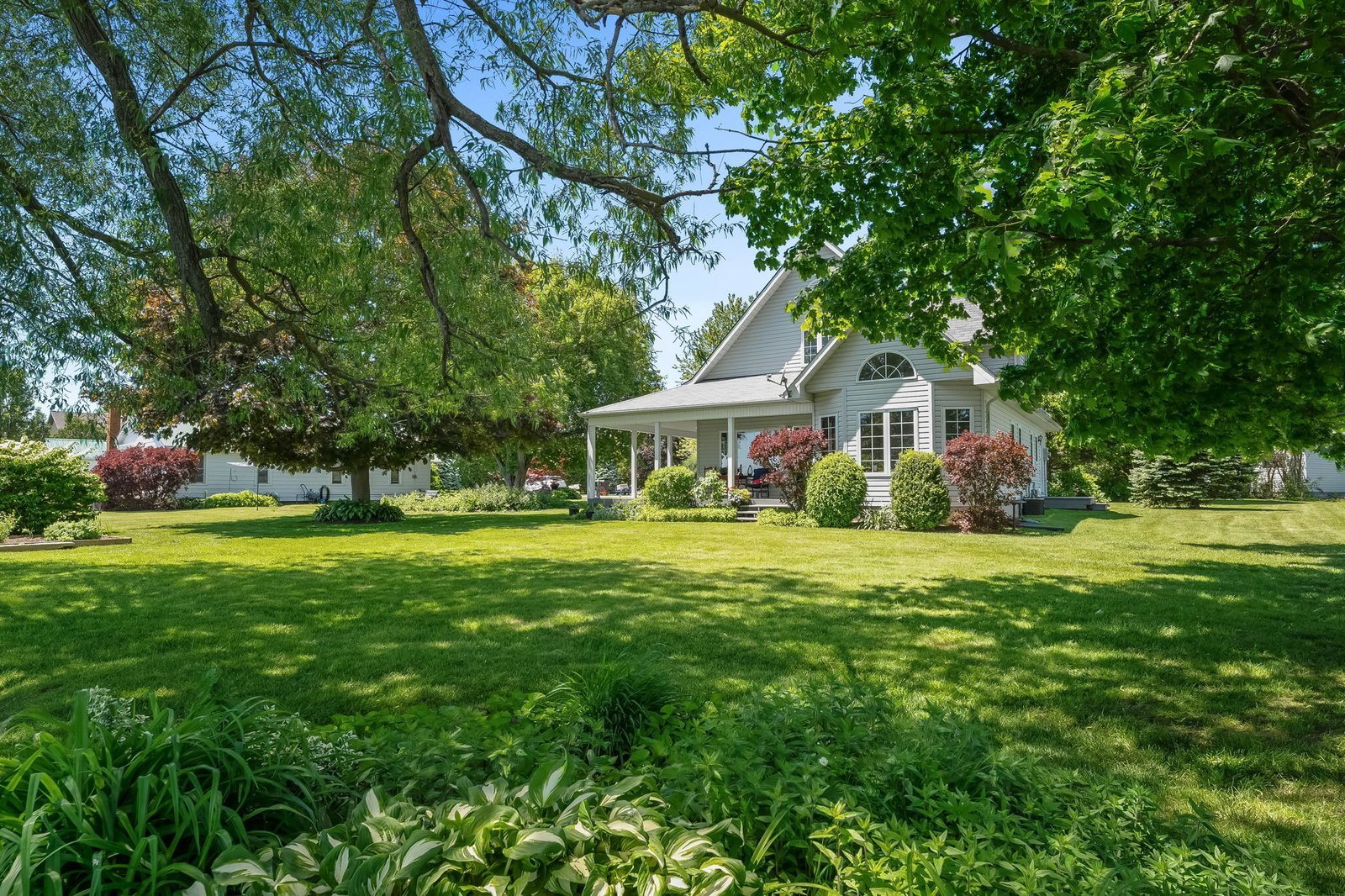 A white house with a large porch sits on a spacious, well-manicured lawn surrounded by green trees and shrubs under a clear blue sky.
