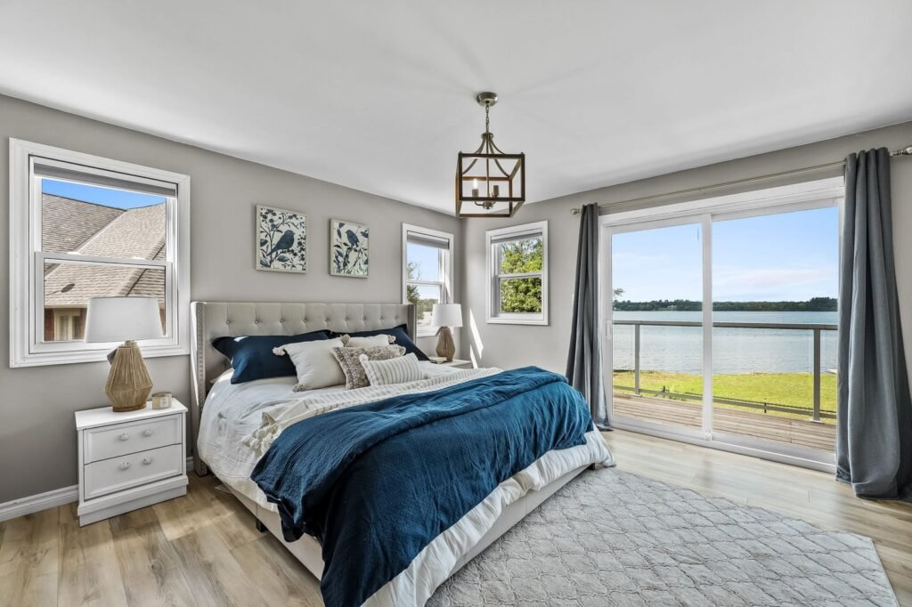 A modern bedroom with a large bed, white and blue bedding, two nightstands, and windows overlooking a lake and balcony.
