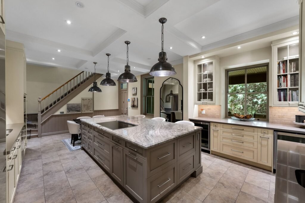Modern kitchen with large central island, pendant lights, beige cabinetry, stainless steel appliances, tiled floor, and staircase leading upstairs.