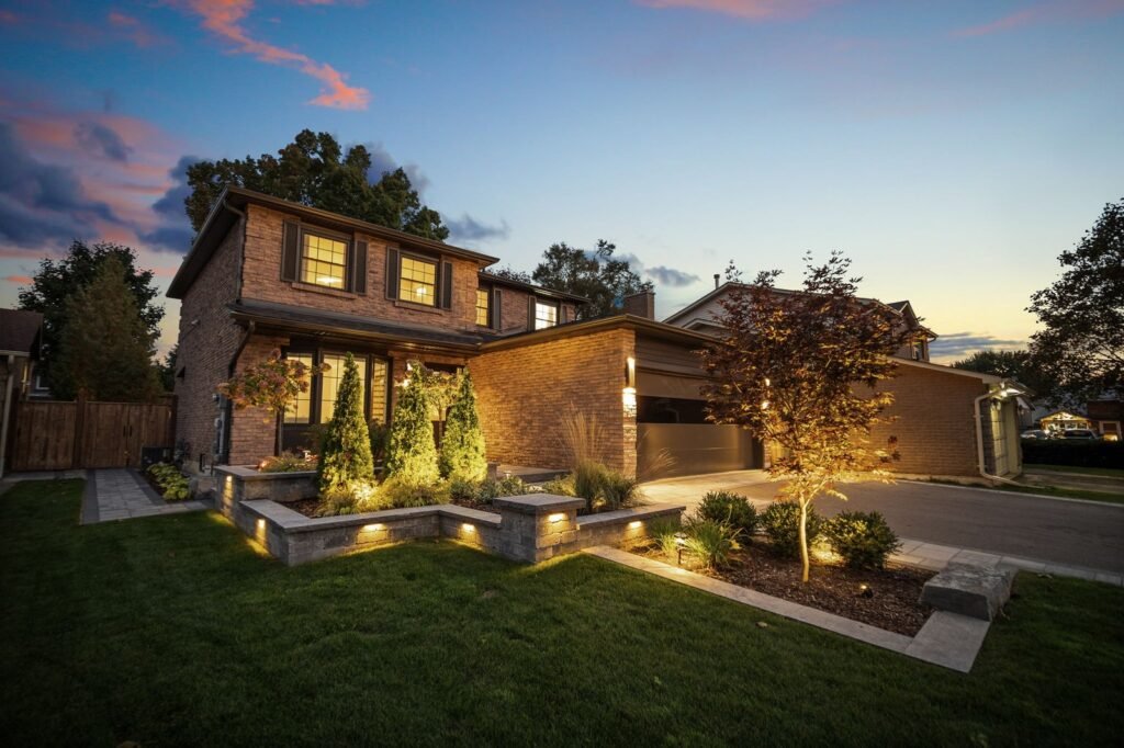 Two-story brick house with large windows, a double garage, lit landscaping, and a manicured lawn at dusk.