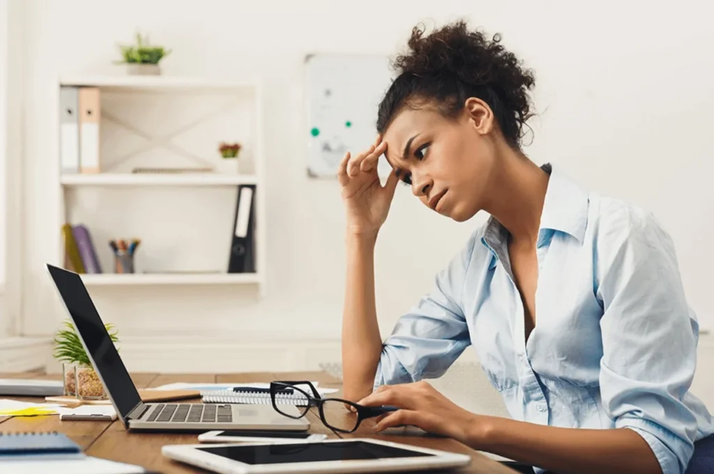 A woman sits at a desk with a laptop, holding her glasses and looking stressed while touching her forehead. Papers and a tablet are also on the desk.