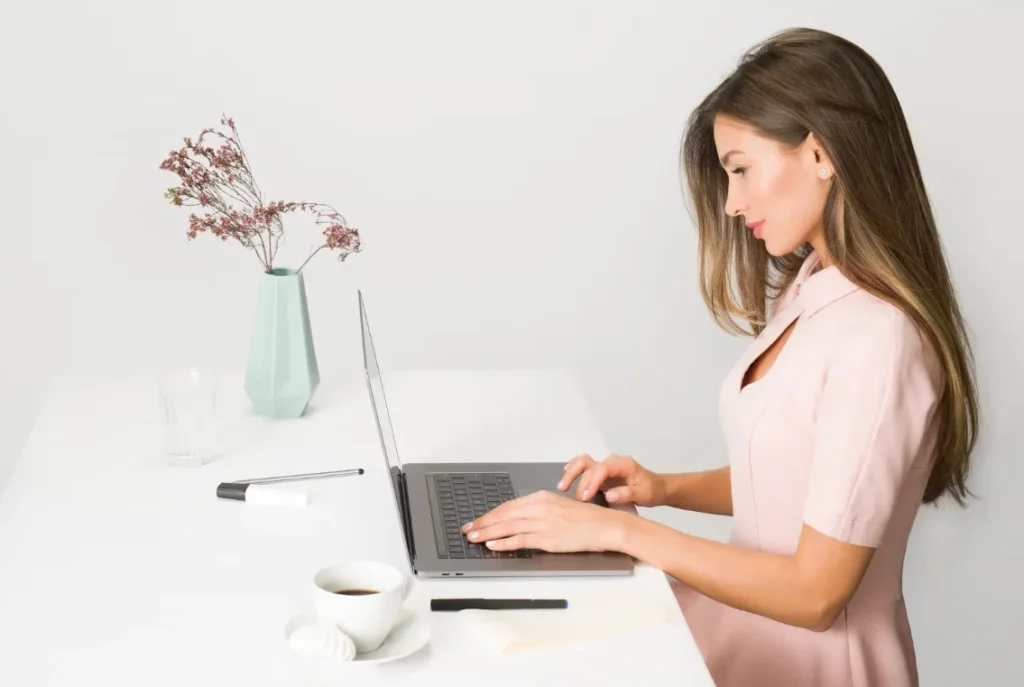 Woman in a pink dress types on a laptop at a white desk with a cup of coffee, a pen, a notepad, and a vase with dried flowers.
