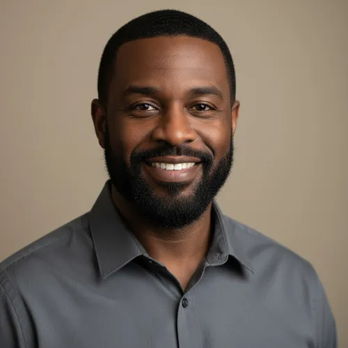 A man with short hair and a beard, wearing a gray button-up shirt, smiles at the camera against a plain beige background.