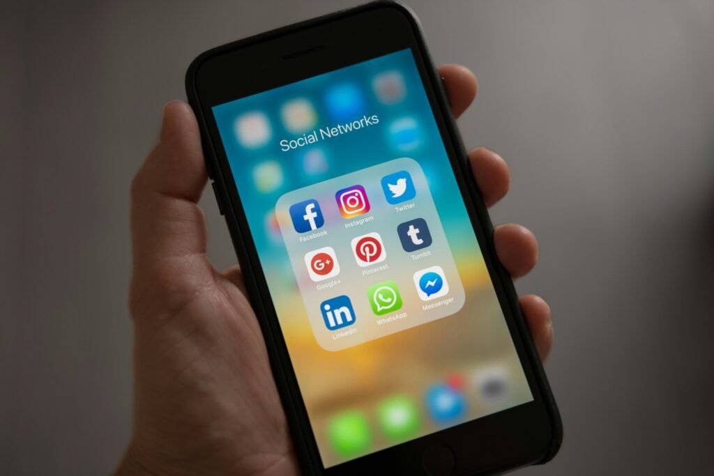 A hand holding a smartphone displaying a folder titled "Social Networks" with app icons for Facebook, Instagram, Twitter, Google+, Pinterest, Tumblr, LinkedIn, WhatsApp, and Messenger.