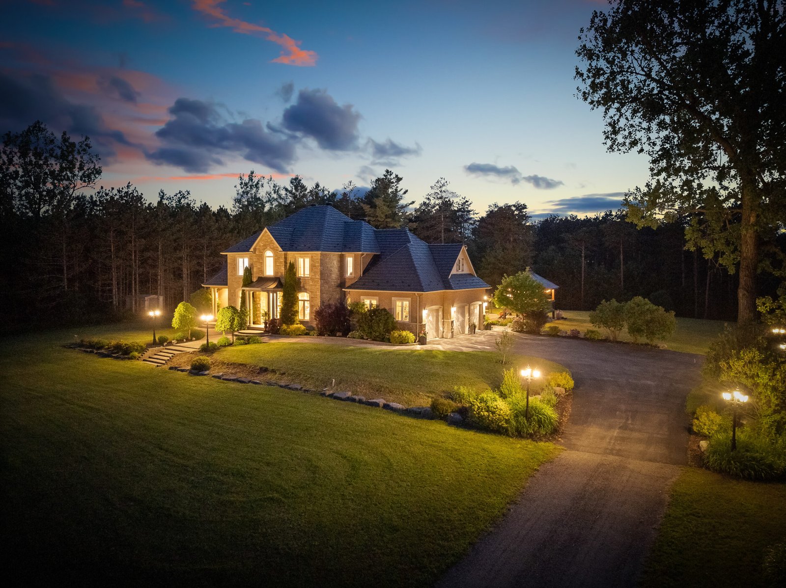 Two-story house with exterior lights on, surrounded by lawn and trees at dusk; paved driveway leads to the main entrance and a detached garage.