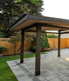 A modern wooden pergola with a flat roof stands on a paved patio in a backyard with a wooden fence and green landscaping.