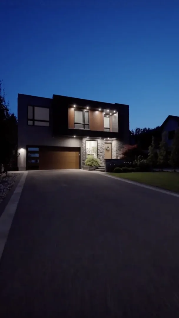 Modern two-story house with large windows and a brown garage, illuminated at dusk, viewed from the driveway.