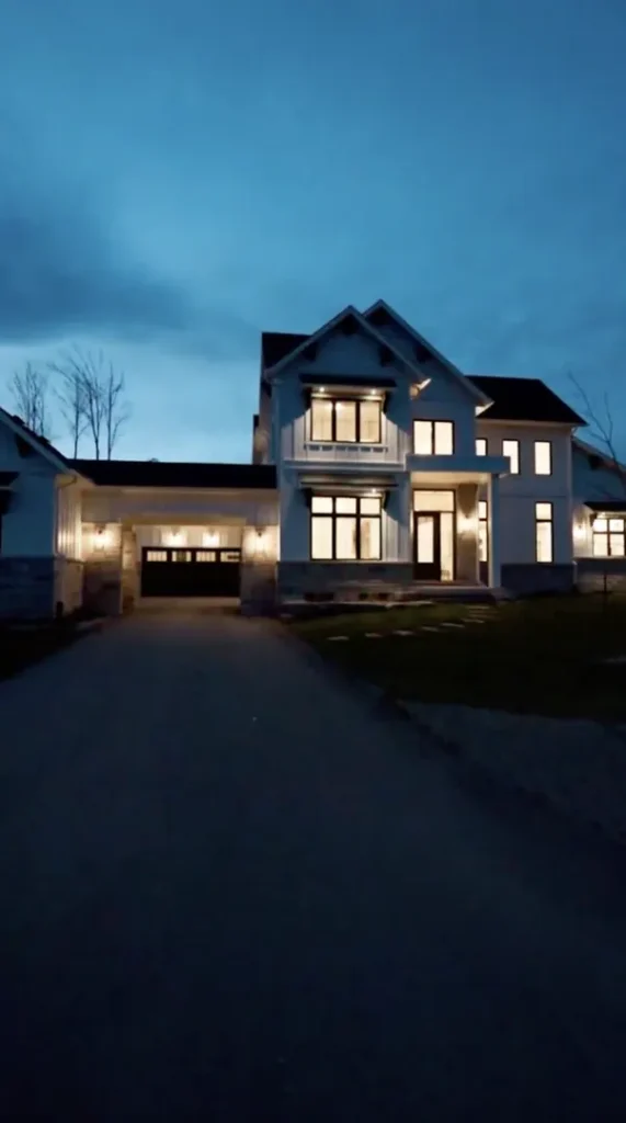 Two-story modern house with illuminated windows and attached garage, photographed at dusk with a dark driveway leading up to the entrance.