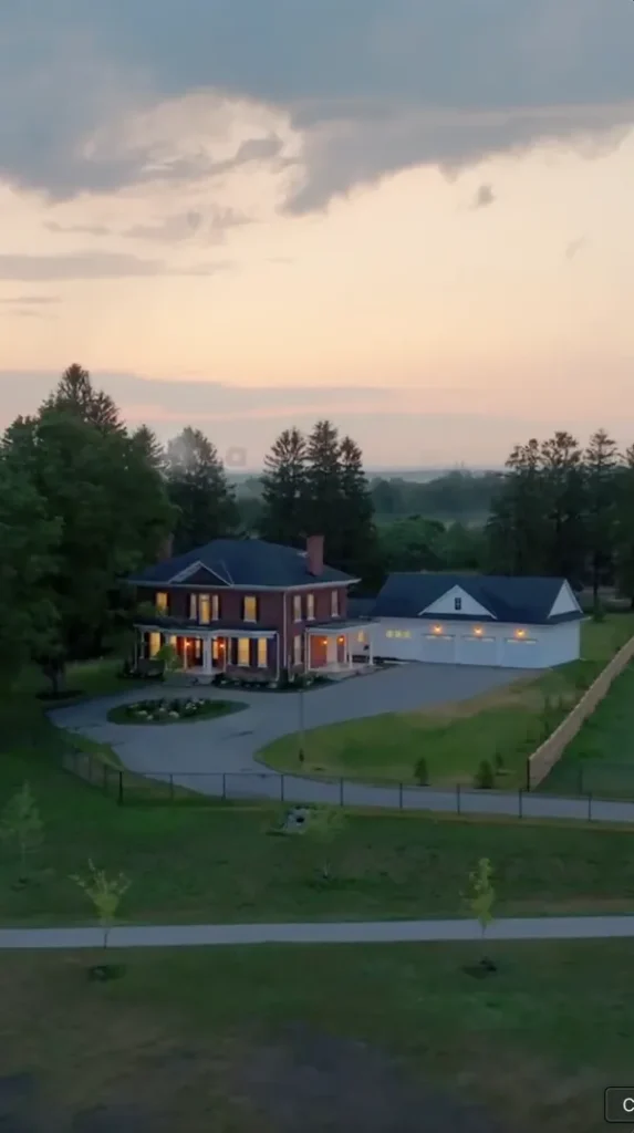 Two-story brick house with white columns and a large driveway, adjacent to a detached garage, surrounded by trees and a fenced yard at dusk.