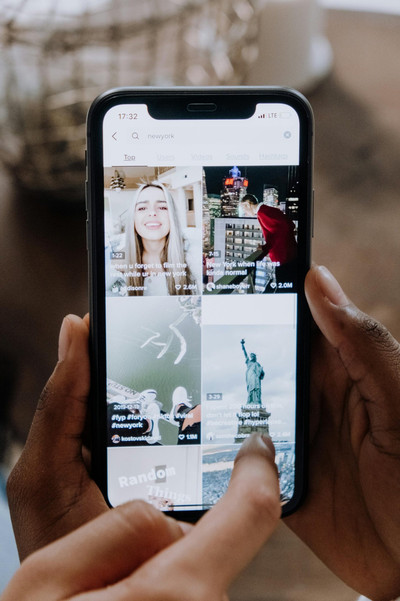 A person holds a smartphone displaying a social media app with four video thumbnails, including a woman, city scenes, and the Statue of Liberty.
