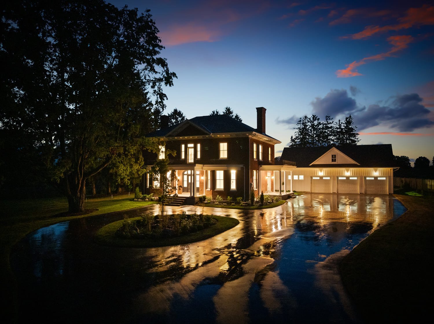 Large two-story house with columns and attached garage, exterior lights on at dusk, reflecting on wet driveway with a tree and landscaped yard.