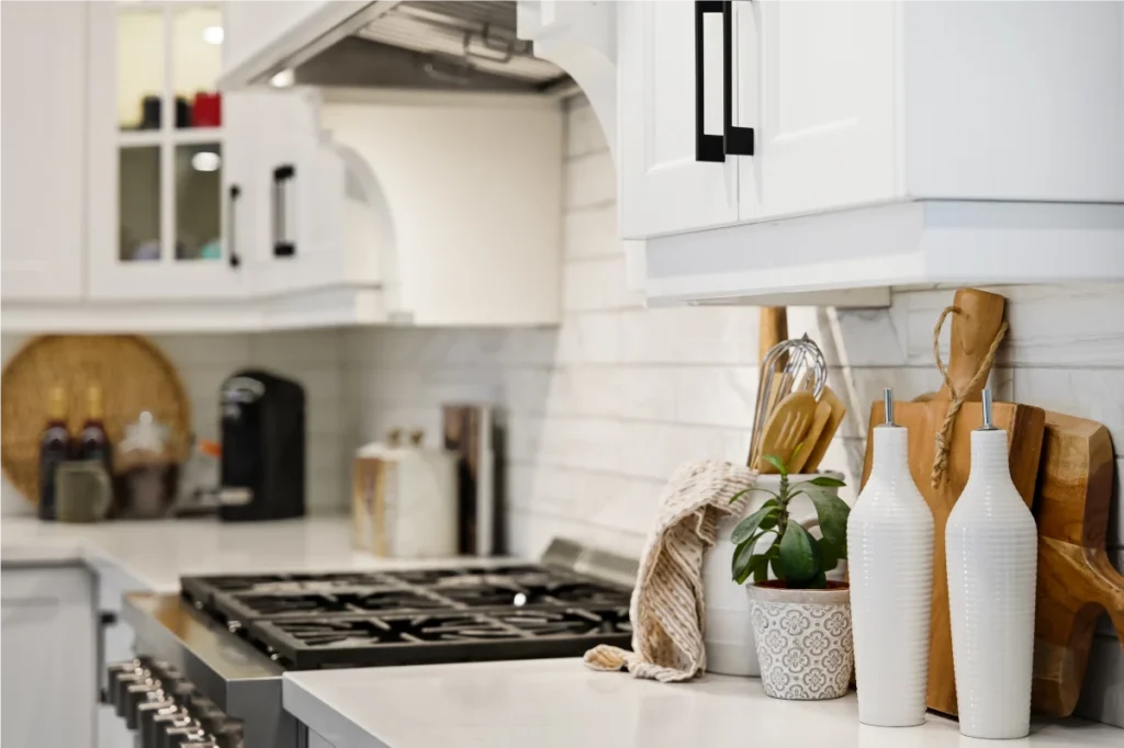 A modern kitchen countertop with a gas stove, white cabinets, cutting boards, a plant, two white bottles, utensils, and a coffee maker.