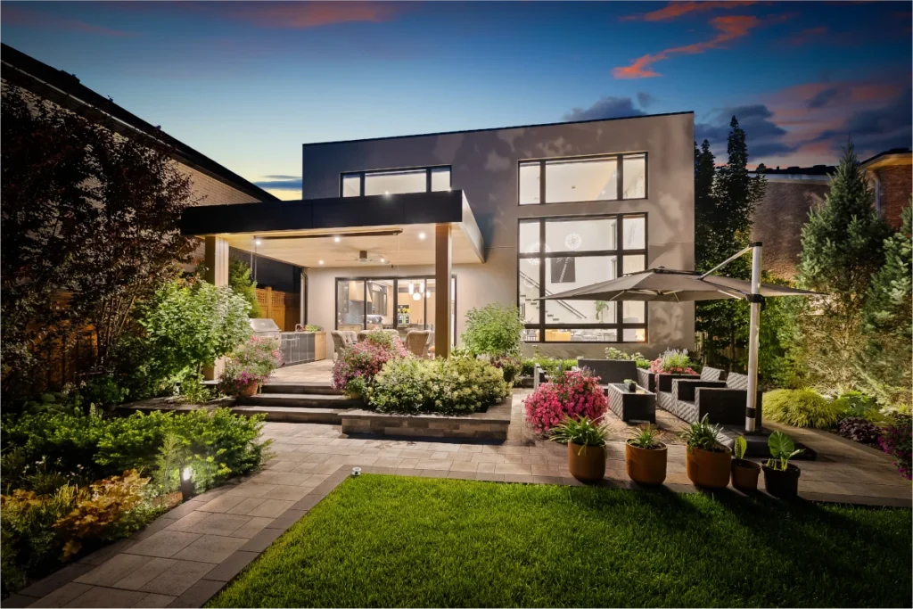 Modern house with large windows, a covered patio, outdoor seating area, lush landscaped garden, and evening lighting.