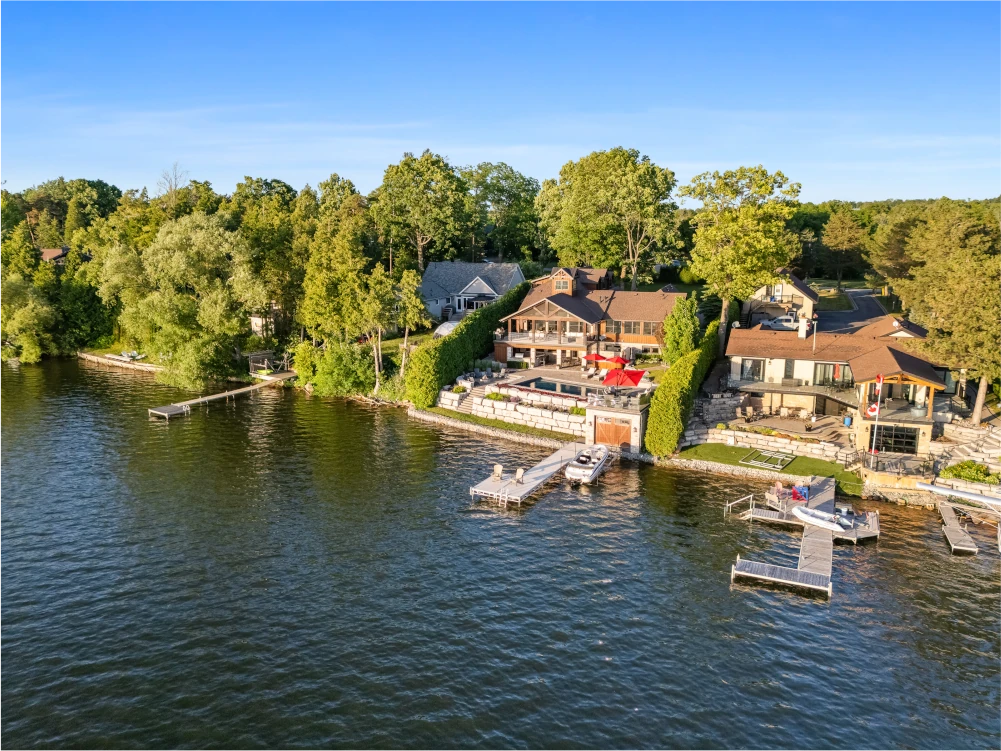 Aerial view of lakeside houses with docks, patios, and boats along a tree-lined shoreline under a clear blue sky.