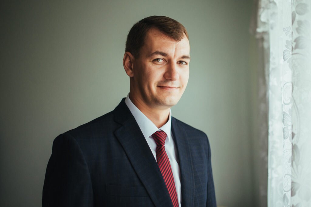 A man in a dark suit and red tie stands indoors near a window with white lace curtains, looking at the camera with a slight smile.